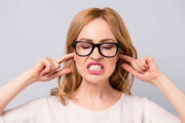 Angry pretty young woman in spectacles covering her ears with fingers and showing teeth