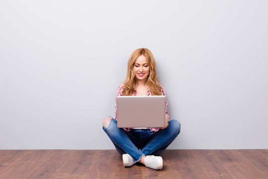 Happy young cheerful woman in checkered shirt and jeans sitting on the floor with laptop on her knees and typing email to a friend