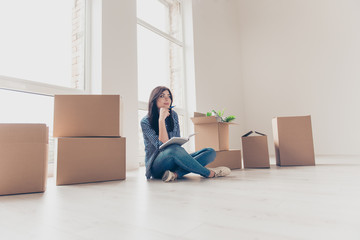 Pretty woman moved in to new house. Smiling young happy woman sitting on the floor surrounded by boxes with stuff writing her ideas about interior to nodepad in her hands