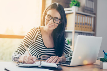 Portrait of smiling woman in glasses making some notes in her diary connected with her work. She...