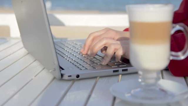 Young woman typing on her laptop in cafe on the beach with glass of latte