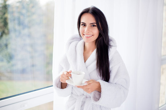 Nice beginning of day! Portrait of gorgeous brunette girl in bathrobe standing near window. She is smiling and holding a cup of coffee