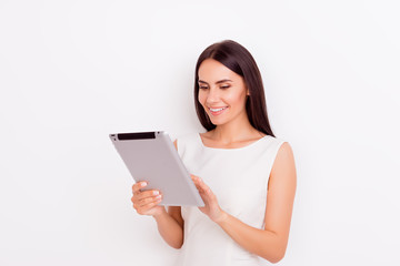 Fototapeta premium Gorgeous young brunette is holding laptop and browsing info on it. She is smiling, wearing white outfit and behind her is a pure background