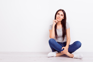 Fototapeta na wymiar Portrait of attractive dreamy pensive brunette lady. She is wearing casual outfit and sitting on the floor with crossed legs on pure white background