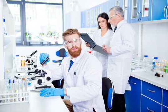 Portrait of young handsome bearded stylish intern, who is wearing gloves and labcoat, safety glasses. Behind him are people who are working for same experiment