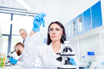 Young brunette cute lab worker in safety glasses is analysing the sample in the lab tube. She is in...