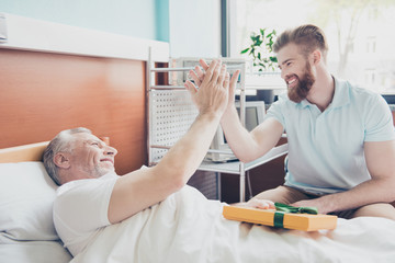 Give me five! Young stylish red bearded guy is visiting and cheering up his grandpa lying in bed at hospital ward