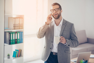 Happy handsome businessman in white shirt and jacket talks on the cell phone in office while drinking coffee