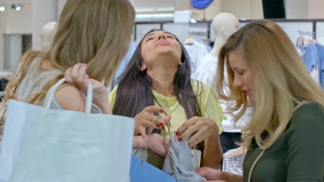 Medium shot of laughing Hispanic woman and her Caucasian female friends with blond hair looking at purchased blouse and chatting with excitement 