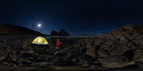 Spherical panorama 360 180 of man at tent on stone beach on shore of Lake Baikal at night