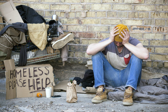 Sad homeless man with  cardboard, sits near  wall. Carriage is home