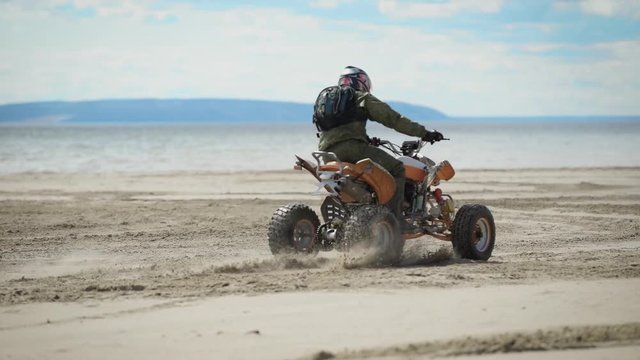 River bank. Sunny weather on the beach. A man is riding an ATV on fine sand. The racer on the ATV carries out roundabouts.