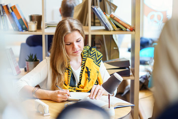 One european young attractive woman is reading book and write some thing in notebook by pen on the table inside light cafe at day time