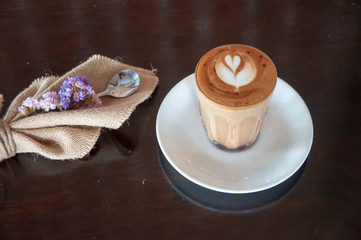 A hot coffee with foam arts in a glass on a woods table. Flowers and fabric decoration.