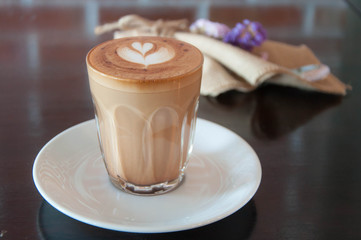 A hot coffee with foam arts in a glass on a woods table. Flowers and fabric decoration.