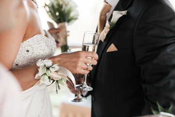 Happy newlyweds couple drink white wedding champagne wine.Decorated crystal glasses.Hands of bride and groom with gold ring toast with alcohol drinks.New family,husband and wife celebrate engagement