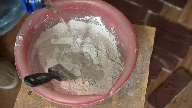 The man pours water into a pink plastic pelvis with a gray powder, then starts to mix the putty with a trowel, close-up shot.