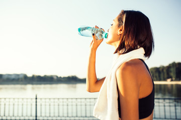 Smiling jogger woman running by the lake at sunset drinks water from a bottle
