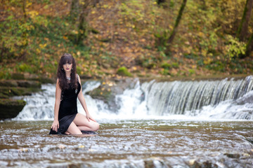 Portrait of a brunette in a black dress near a mountain river. The model is posing at the waterfall.
