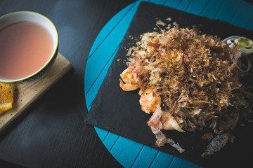 Buckwheat noodles with shrimps and shavings of tuna with sauce on black plate. Asian food background. Eating concept. Restaurant place with wooden table. Flare copy space for text, design