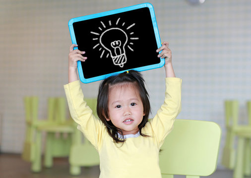 Cute little child girl holding blackboard showing bulbs image in kids room. Education concept.