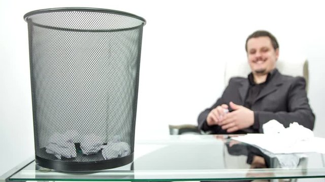 A young man is sitting in his office and is bored. A waste bin is on his desk and he tries to throw crumpled paper into the waste bin and is successful four times in a row.