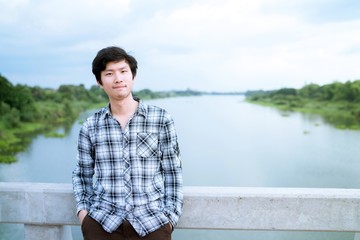 Fototapeta premium Portrait of an interesting asian man with friendly smile in casual wear. Dark Haired Young Man on the river Bridge with copy space.