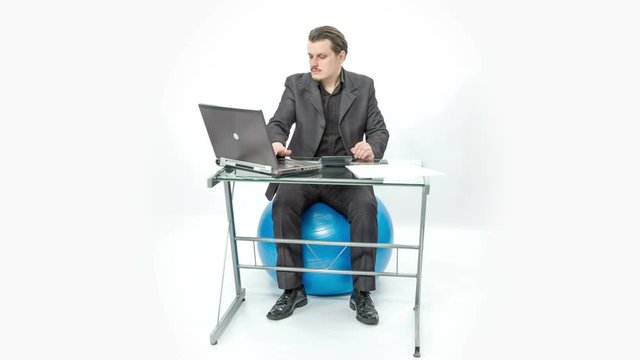 A young man is moving his feet alternately. He is sitting on an exercise ball and he is working at his desk in the office.