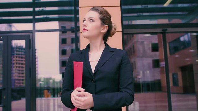 Attractive young business woman is waiting for a meeting. Girl in business clothes with a red folder in hands on the background of a business center. SLOW MOTION