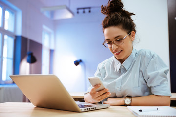Young beautiful woman wearing glasses working in the office, uses a laptop and mobile phone