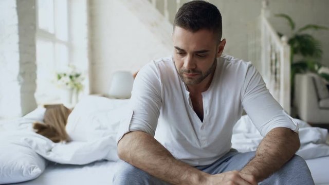 Upset young man sitting in bed have problems with sex while his girlfriend sleep in bedroom at home