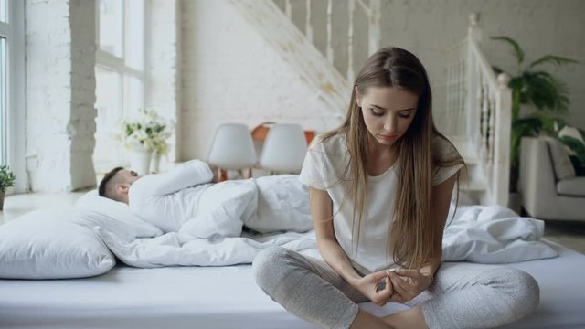 Depressed young woman sitting in bed and crying while her boylfriend lying in bed at home
