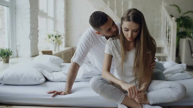 Depressed young woman sitting in bed and crying while her boylfriend come and embrace her and kiss in bedroom at home