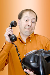 A white man in an orange shirt and glasses with a telephone receiver in the rivers looks at the old disk-type wired telephone with surprise and bewilderment