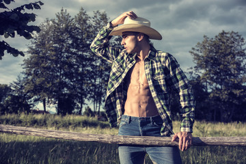 Portrait of sexy farmer or cowboy in hat with unbuttoned shirt on muscular torso, looking to a...