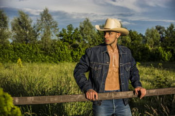 Portrait of sexy farmer or cowboy in hat with unbuttoned shirt on muscular torso, looking to a side, while standing next to hay field in countryside