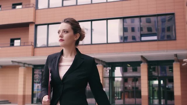 Beautiful confident young business woman is in a hurry to negotiate. Girl in business clothes with a red folder in hands on the background of an office building