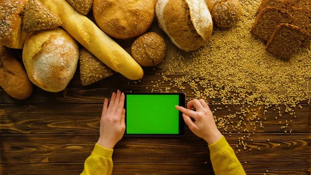 Woman using tablet device with green screen on wooden table with fresh bread. Bakery background. Fresh whole grain bread and buns. Top view. Female hands scrolling pages, tapping on touch screen