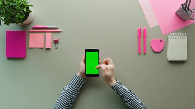 Top view woman using smart phone with green screen on the grey table background with pink details. Woman holding cell phone in hands and swiping, scrolling pages. Close up. Above shot. Chroma Key