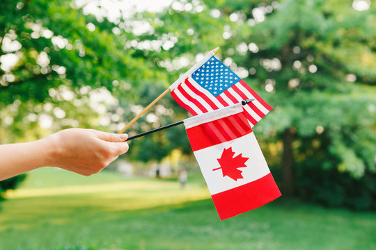 Macro Closeup Shot Of Hand Arm Holding Waving Canadian And American Flags On Green Forest Park Nature Background Outside, Celebration Of National Or Flag Day Concept