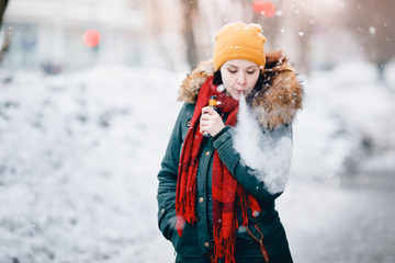 Fototapeta premium girl is holding an electronic cigarette in her hand and exhaling steam from her mouth. She is wearing a hat. Winter. Concept of safe smoking, giving up tobacco. Toning, highlighting