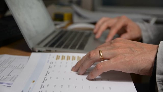 Hands of a man working, reading data on a paper sheet and using portable computer typing on keyboard in his office