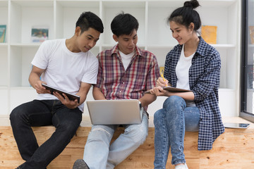 Fototapeta premium young asian people are using different gadgets and smiling, sitting near white wall. Male and female students studying using laptop computer, tablet.