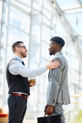 Portrait of smiling businessman chatting with African-American partner patting him on shoulder in modern glass hall of office building