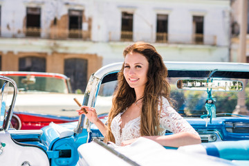 Beautiful young bride in white wedding dress is sitting in blue retro car cabriolet with cigar in her arm