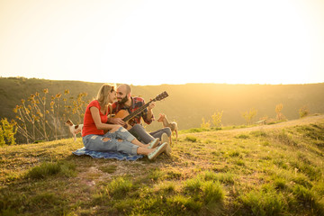 Fototapeta na wymiar Lovers in nature. Young couple in love sitting on the park while these young guitar playing guitar in sunset time. Together with dogs