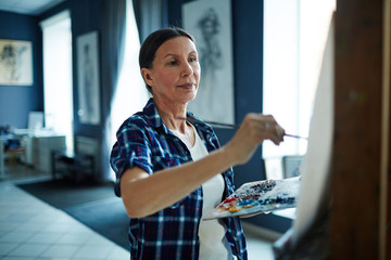 Obraz premium Portrait of elegant mature woman painting oil picture on canvas in art-studio, looking inspired and enjoying process