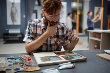 Portrait of contemporary bearded artist working in art studio painting pictures thinking and looking focused