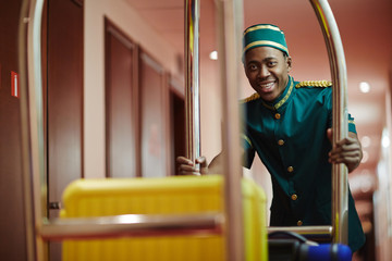 Portrait of smiling African bellhop helping guests, pushing luggage cart delivering bags to hotel...