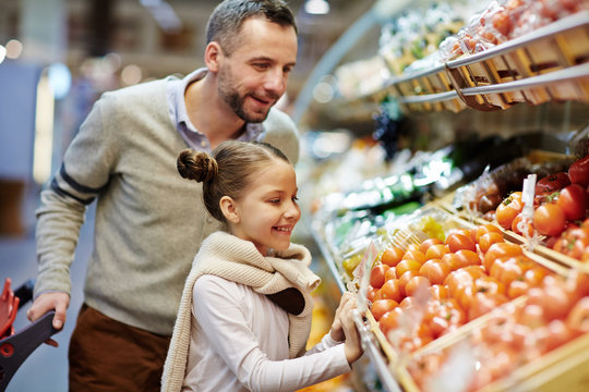 Portrait of little girl with dad leaning over vegetable counter choosing fresh ripe tomatoes and other vegetables in supermarket